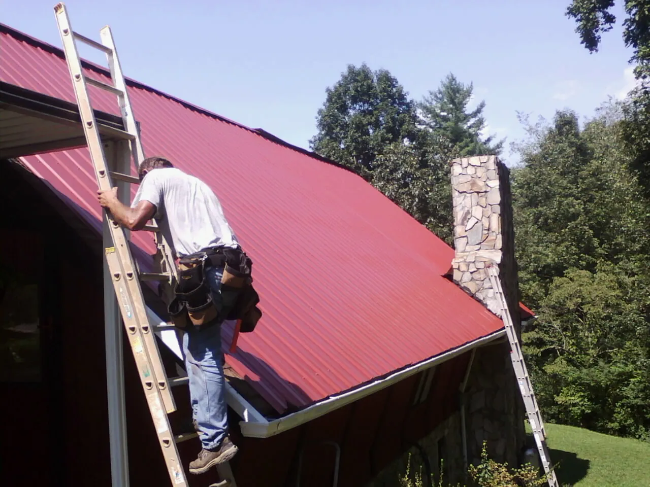 Roofer on a ladder beside a red metal roof and stone chimney — Soffit & Fascia Repair by Roofing Brothers in Moshannon